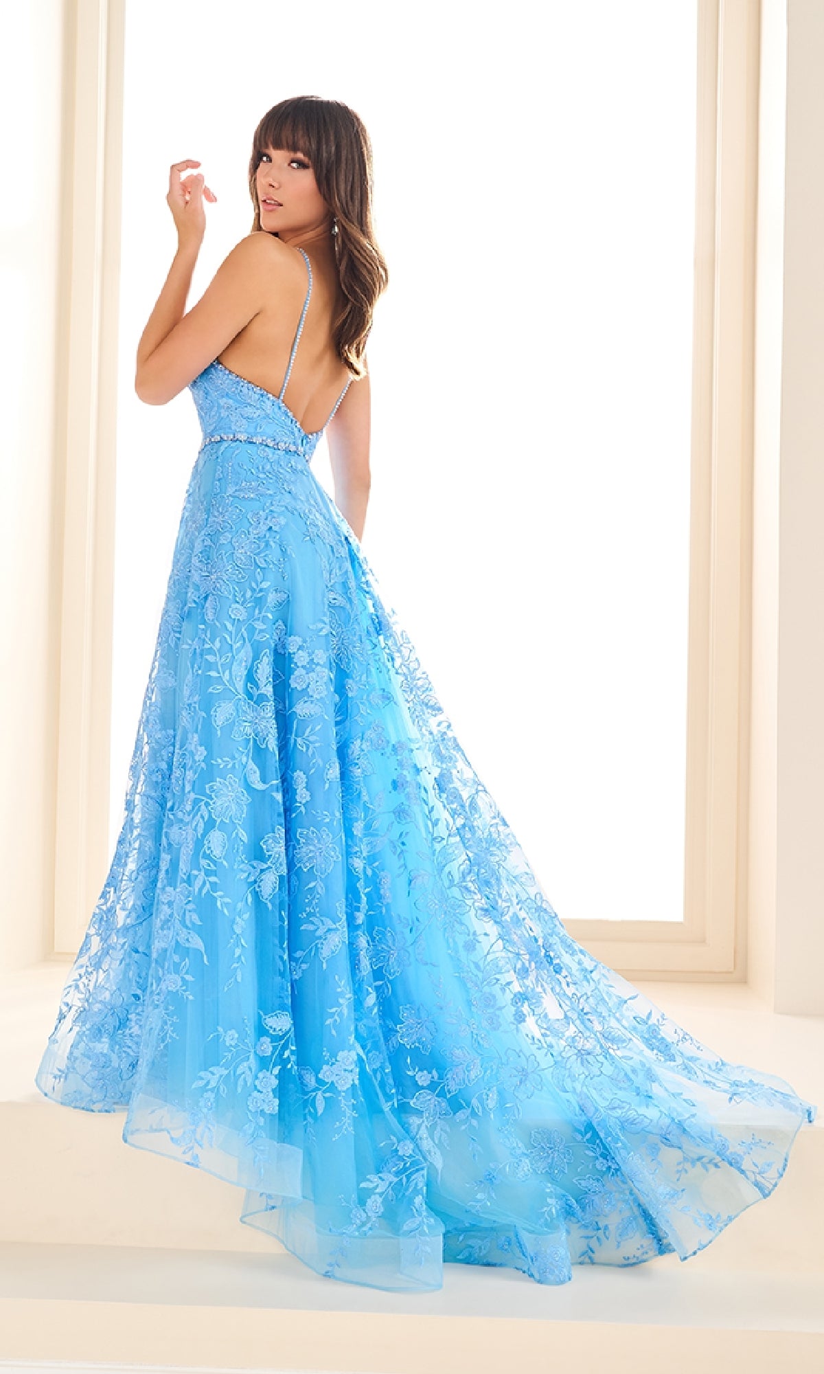 A woman in the Ellie Wilde EW36088 long prom dress—light blue embroidered tulle, a-line skirt with pockets, and spaghetti straps—stands looking over her shoulder in a well-lit room.